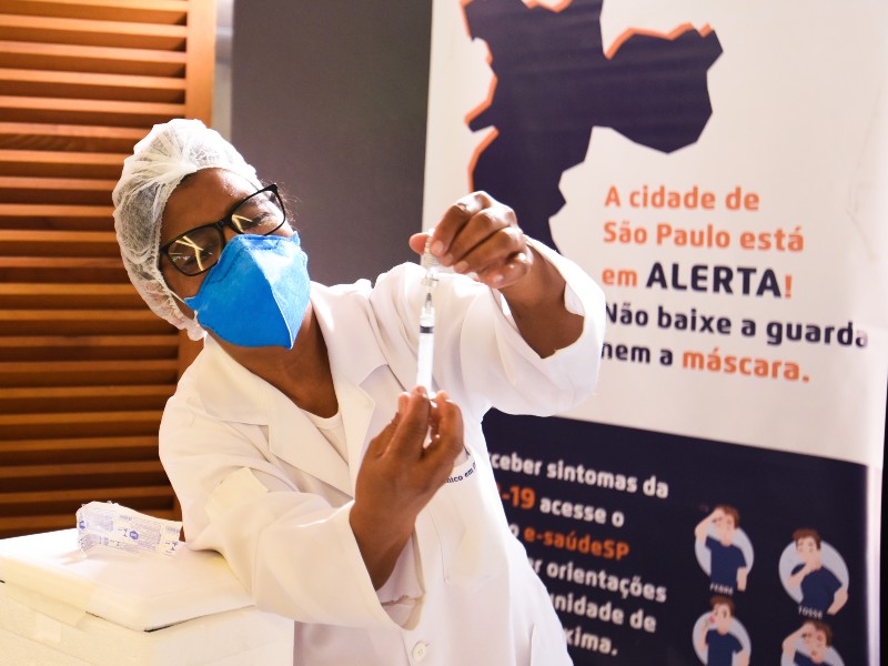 Foto de uma profissional da saúde manipulando vacina. Com uma das mãos ela aspira a vacina com a seringa enquanto segura o frasco com a outra mão, olhando atentamente para o procedimento. Do lado direito está um cartaz com dizeres sobre a importância da máscara e sobre os sintomas da Covid-19